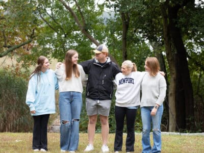 Five teens standing in a line looking at each other with their arms over each others' shoulders.