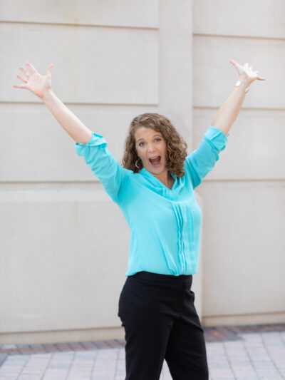 Sam Straub wearing a turquoise shirt and black pants with her arms in the air and mouth wide open smiling in celebration