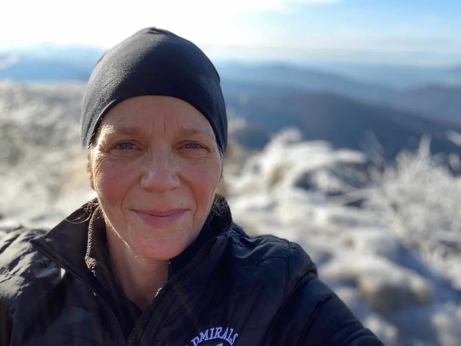 IMG_9600 Close up of Sam Straub wearing a black hat on a mountain top with ice covered branches and mountains in the background.