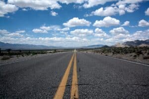 Yellow lines on a concrete road leading off to a horizon line with mountains and a blue sky filled with white clouds