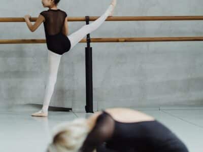 Two teen ballet dancers stretching. One is in profile, closer to the camera sitting on the floor in a straddle.  The other has her back to the camera with her leg up on a bar.
