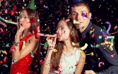 Three teens with party blowers in their mouths and confetti in the air all around them. The boy is looking at the camera. Two girls are looking off to the left