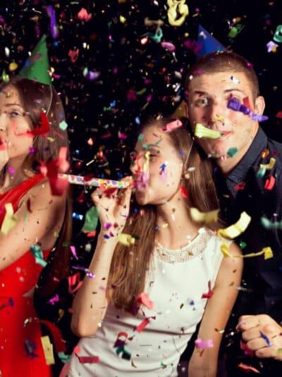 Three teens with party blowers in their mouths and confetti in the air all around them. The boy is looking at the camera. Two girls are looking off to the left