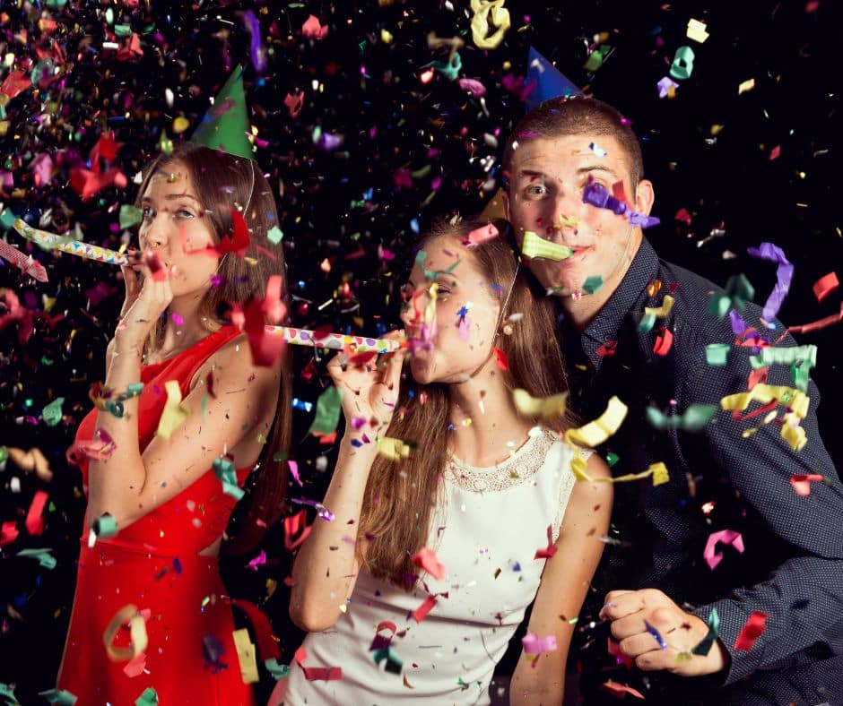 1 Three teens with party blowers in their mouths and confetti in the air all around them. The boy is looking at the camera. Two girls are looking off to the left