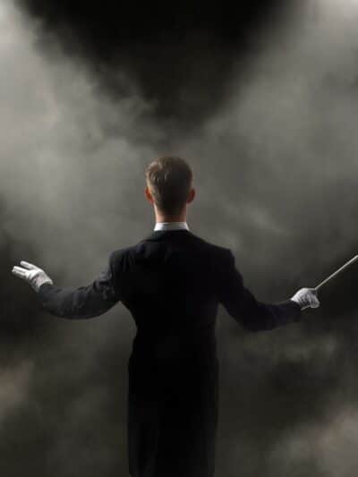 View of an orchestra conductor from the back. Holding a baton in right hand. Wearing white gloves and a black tux with a white color. In front of the male conductor are two spot lights shining through smoke.