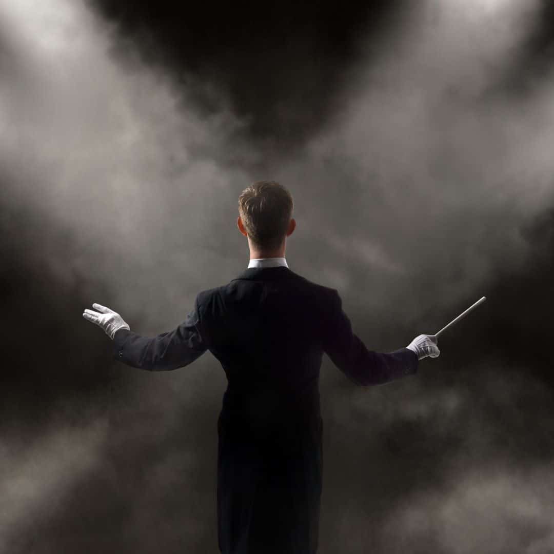 View of an orchestra conductor from the back. Holding a baton in right hand. Wearing white gloves and a black tux with a white color. In front of the male conductor are two spot lights shining through smoke.
