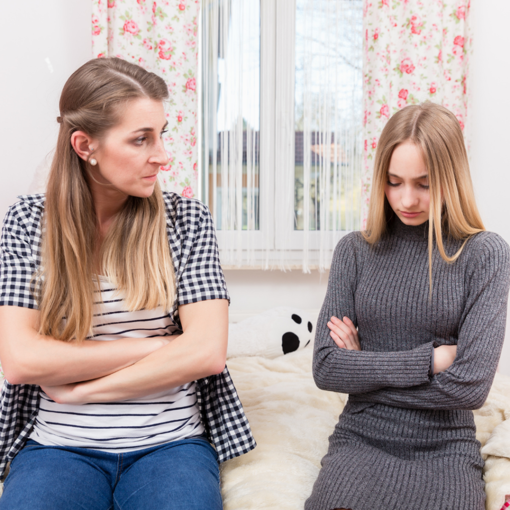 Mom and teen daughter facing the camera with their arms folded.  Mom is looking at daughter disapprovingly. Daughter is looking down at the floor.