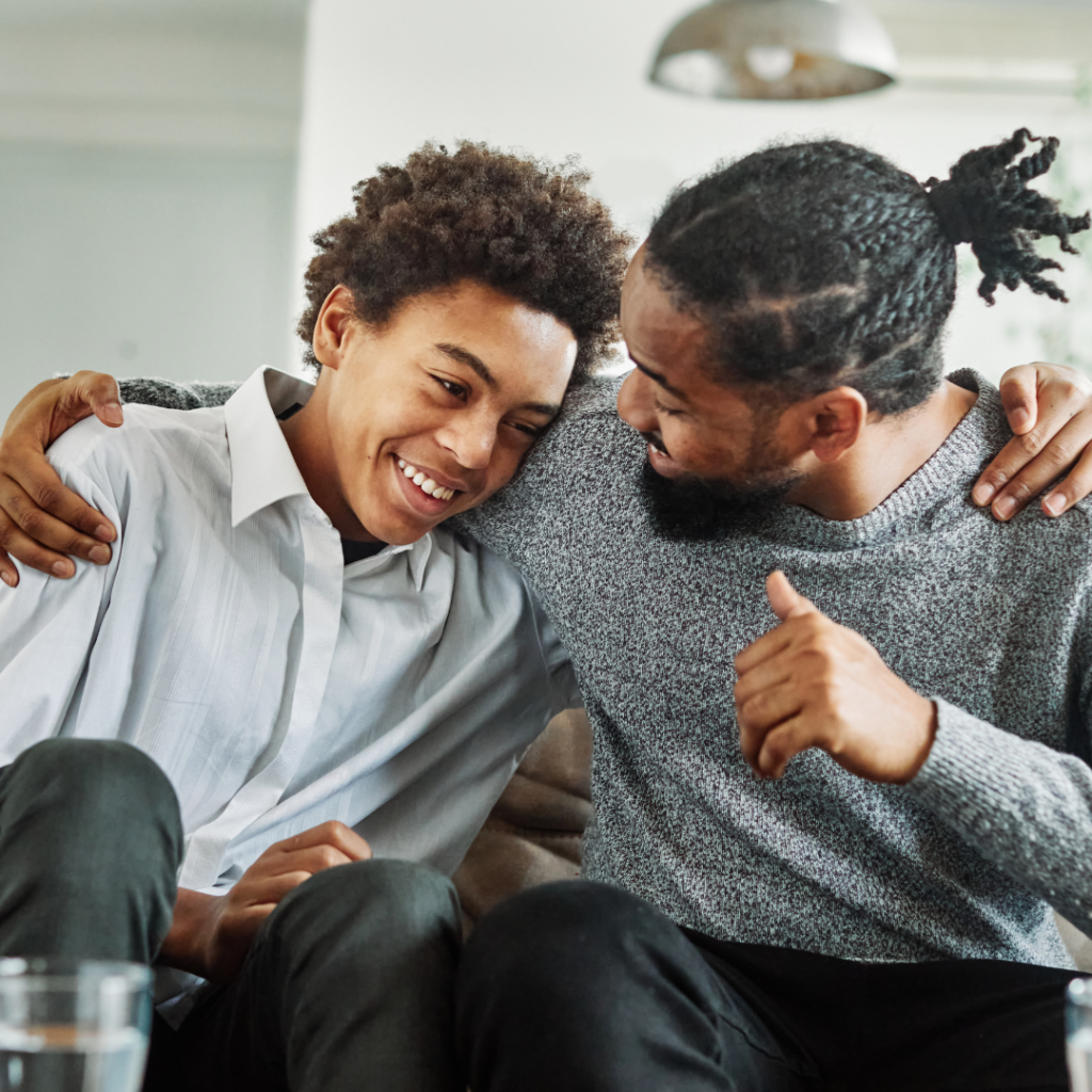 Dad with his arm around his teenage son.  Son is smiling and leaning into Dad's shoulder. Both are sitting on a couch. 