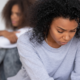 Mom looking looking upset with her head down close to the camera and with back to daughter. Teen daughter in the background with arms folded, not looking at Mom.