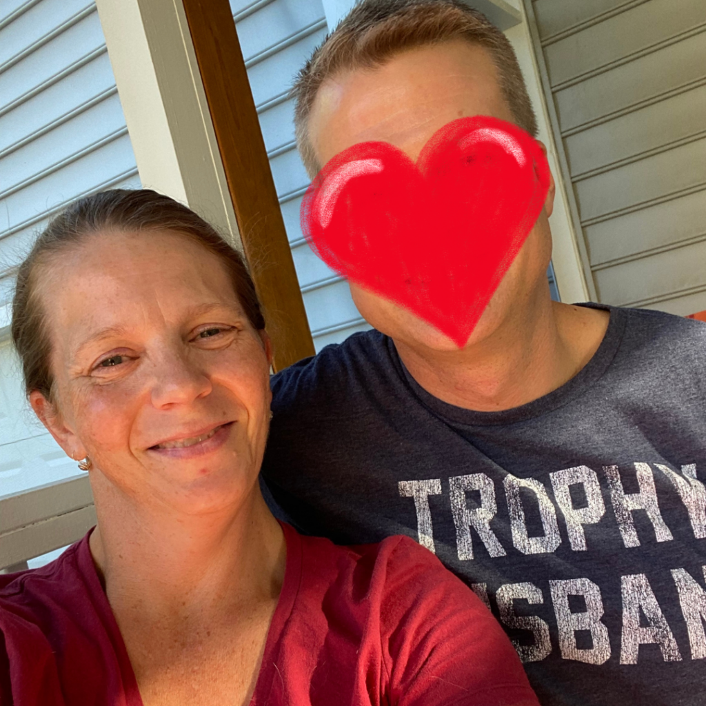 Sam Straub and her husband posing for a selfie.  Husband is wearing a t-shirt that says "Trophy Husband" and a heart image has been placed over his face.