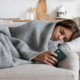 Teen girl wrapped in a grey blanket lying on a couch looking at a cell phone. Kitchen wall in the background