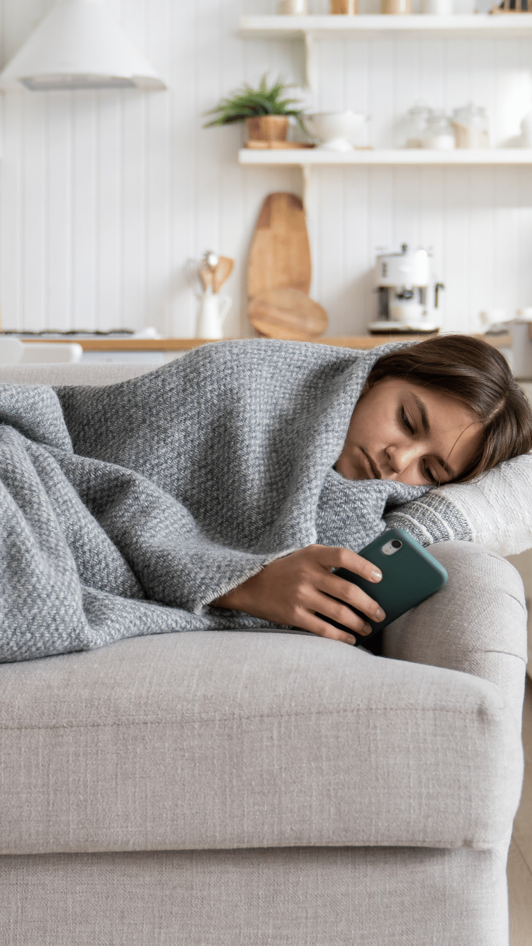 Untitled (Your Story) Teen girl wrapped in a grey blanket lying on a couch looking at a cell phone. Kitchen wall in the background